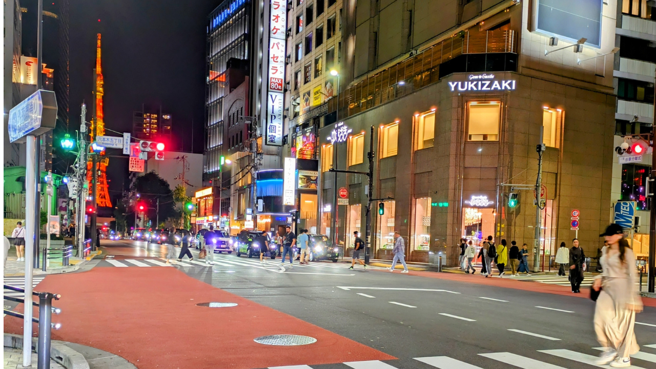 City street at night with a lit red-and-white tower in the distance and people crossing